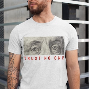 Może przedstawiać: Szary T-shirt z zbliżeniem oczu Benjamina Franklina z amerykańskiego banknotu dolara. Tekst "TRUST NO ONE" jest nadrukowany na czerwono pod obrazkiem.