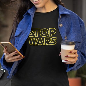 May include: A person wearing a black t-shirt with the text "STOP WARS" in yellow outlined letters. The person is wearing a blue denim jacket and holding a smartphone and a cup of coffee.