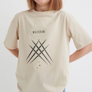 May include: A beige t-shirt featuring a black graphic of Wolverine's claws dripping with blood. The text "Wolverine" is printed above the graphic.