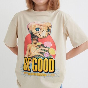 Pode incluir: Uma camiseta bege com uma estampa gr&aacute;fica de um personagem alien&iacute;gena fict&iacute;cio segurando flores. O texto "BE GOOD" est&aacute; impresso em letras amarelas abaixo do personagem. O texto "THE EXTRA TERRESTRIAL" est&aacute; impresso em letras menores abaixo do texto principal.