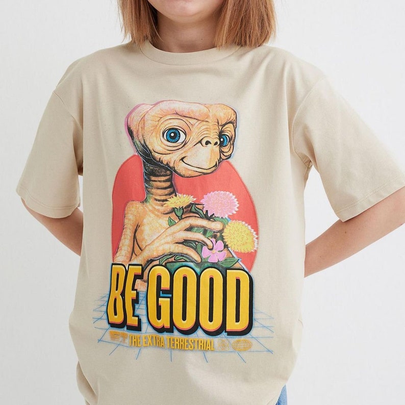 Pode incluir: Uma camiseta bege com uma estampa gr&aacute;fica de um personagem alien&iacute;gena fict&iacute;cio segurando flores. O texto "BE GOOD" est&aacute; impresso em letras amarelas abaixo do personagem. O texto "THE EXTRA TERRESTRIAL" est&aacute; impresso em letras menores abaixo do texto principal.