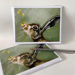 May include: A set of blank greeting cards featuring a painting of a brown and white hare sniffing a yellow flower. The hare has large ears and is looking up at the flower.
