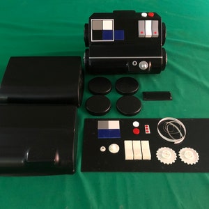 Puede incluir: Un conjunto de piezas de plástico negro para un kit de maquetas. El conjunto incluye un cuerpo principal, dos paneles laterales, cuatro piezas redondas, una pieza rectangular pequeña, un panel con un cuadrado azul y blanco, tres botones rojos, un botón blanco, dos piezas rectangulares blancas, dos engranajes blancos y una cuerda de plástico transparente.