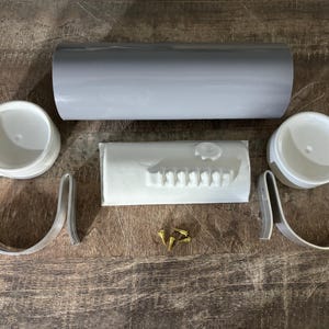 Op de afbeelding: Een set witte kunststof onderdelen voor een gootsteensysteem, inclusief twee gebogen beugels, twee eindkappen, een recht stuk en een rij tanden. De onderdelen liggen op een houten oppervlak met een grijze buis en kleine gouden schroeven.