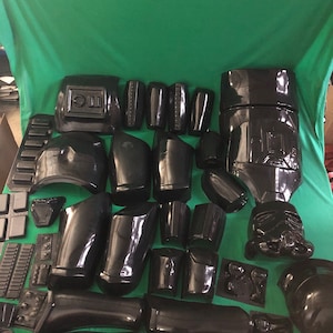 May include: A set of black plastic pieces for a costume. The pieces are arranged on a green surface.