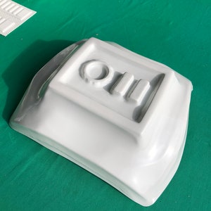 May include: A white ceramic dish with a raised design of a circle and three rectangles. The dish is on a green surface.