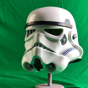 Könnte beinhalten: Weißer Stormtrooper-Helm mit schwarzen und blauen Details. Der Helm hat eine glatte, glänzende Oberfläche und weist ikonische Gesichtsdesignelemente auf, darunter ein schwarzes Visier und Belüftungsmerkmale. Der Helm wird auf einem Ständer vor einem grünen Hintergrund ausgestellt.