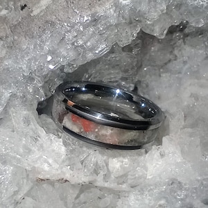 May include: A silver ring with a thin black band and a clear stone inlay with red and white flecks. The ring is resting on a bed of white crystals.