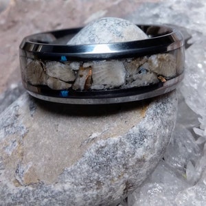 Pu&ograve; includere: Un anello in carburo di tungsteno nero con una fascia di materiale fossilizzato. L'anello ha una finitura lucida e un bordo smussato.