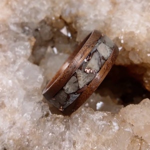 Pu&ograve; includere: Un anello in legno con una fascia di conchiglia fossilizzata e intarsi in rame. L'anello &egrave; posto su uno sfondo di cristalli bianchi.