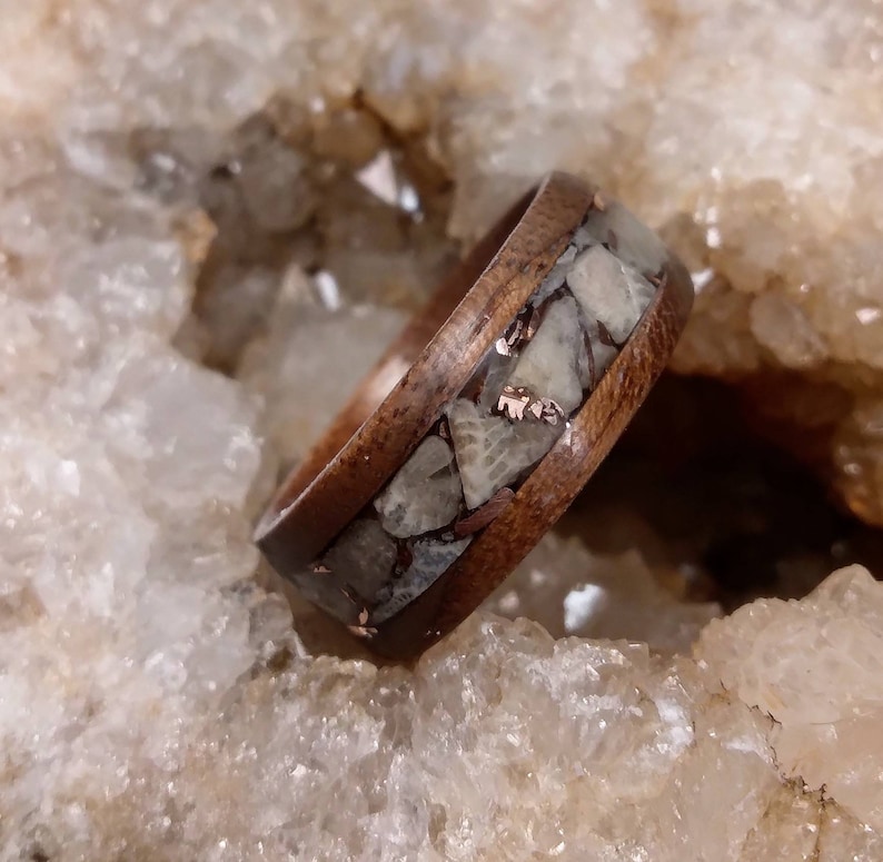 Pu&ograve; includere: Un anello in legno con una fascia di conchiglia fossilizzata e intarsi in rame. L'anello &egrave; posto su uno sfondo di cristalli bianchi.