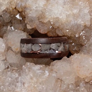 Pu&ograve; includere: Un anello in legno marrone scuro con una sottile fascia di rame e una fascia di cristalli di quarzo bianchi.