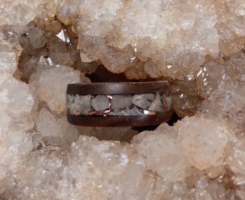 Pu&ograve; includere: Un anello in legno marrone scuro con una sottile fascia di rame e una fascia di cristalli di quarzo bianchi.