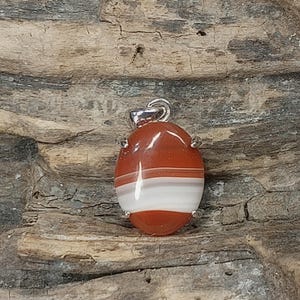May include: An oval-shaped pendant featuring a polished, banded stone with horizontal layers of deep reddish-brown and white. The stone is secured in a silver-toned metal setting with four prongs and includes a small silver-toned metal bail at the top.