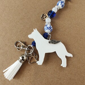 Può includere: Un ciondolo a forma di cane bianco con ali argentate, un ciondolo a forma di zampa e un nappa bianca. Il ciondolo è attaccato a una catena di perline blu e bianche. È incluso anche un ciondolo a forma di zampa dorata.