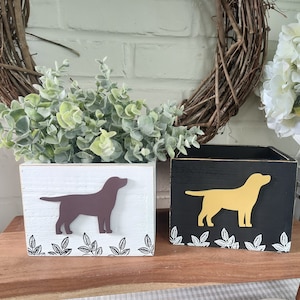 Könnte beinhalten: Zwei Holzkisten, eine weiße und eine schwarze, jeweils mit einer Hunde-Silhouette. Die weiße Kiste hat einen braunen Hund, die schwarze Kiste einen goldenen Hund. Beide haben Blattmuster. Rustikale Dekoration.