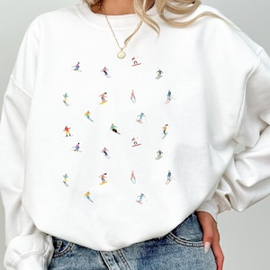 Könnte beinhalten: Weißer Pullover mit einem bunten Grafikdruck von Skifahrern auf weißem Hintergrund. Der Pullover hat einen Rundhalsausschnitt und lange Ärmel.