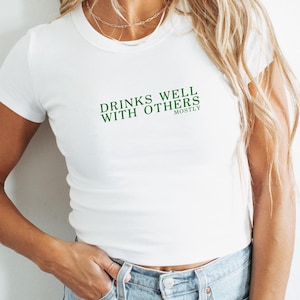 Könnte beinhalten: Weißes, kurzes Baby-T-Shirt mit dem grünen Text "DRINKS WELL WITH OTHERS". Das Wort "mostly" steht unter dem Text. Das Shirt wird mit einer blauen Jeans kombiniert.