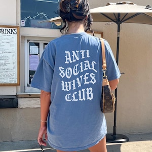 Puede incluir: Una camiseta azul con el texto "Anti Social Wives Club" impreso en blanco en la espalda. La camiseta la lleva puesta una persona que est&aacute; de pie frente a un edificio.