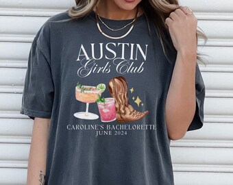 May include: A dark gray t-shirt with the text "Austin Girls Club" and a graphic of two cocktails and a cowboy boot. The text "Caroline's Bachelorette June 2024" is also printed on the shirt.