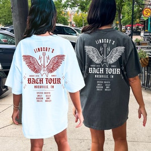 May include: Two light blue and dark gray t-shirts with a graphic print of a guitar with wings. The text on the shirts reads "Lindsay's Bach Tour Nashville, TN".