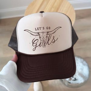May include: A brown and cream trucker hat featuring a foam front panel with a brown graphic of a longhorn skull and the text LET'S GO Girls. The hat includes a brown mesh back, a brown brim with stitching, and a brown cord detail across the base.