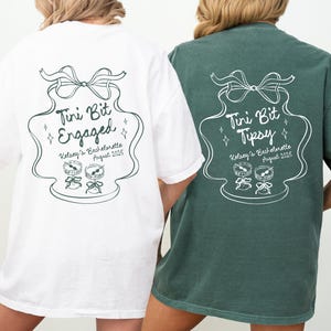 May include: Two white t-shirts with a green design. The design is a stylized illustration of a champagne flute with a bow on top. The text on the shirts reads "Tini Bit Engaged" and "Tini Bit Tipsy".
