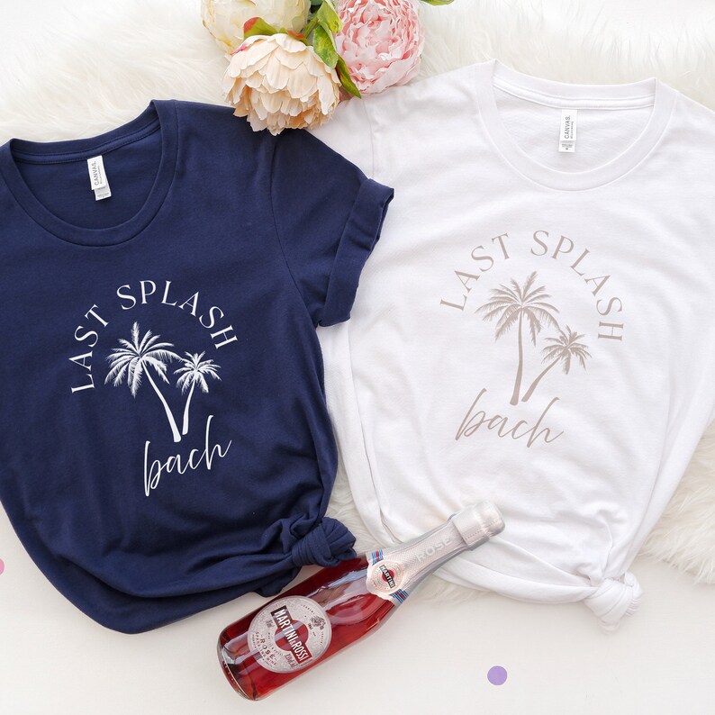 Puede incluir: Dos camisetas blancas con el texto "Last Splash Bach" y palmeras impresas en ellas. Una camiseta es azul marino y la otra es blanca. Una botella de vino espumoso est&aacute; en primer plano.