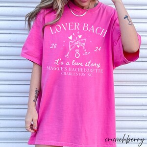 May include: Pink t-shirt with white text that reads "Lover Bach 20 - 24 it's a love story Maggie's Bachelorette Charleston, SC". The shirt features a graphic of two champagne glasses clinking with a ring in the middle.