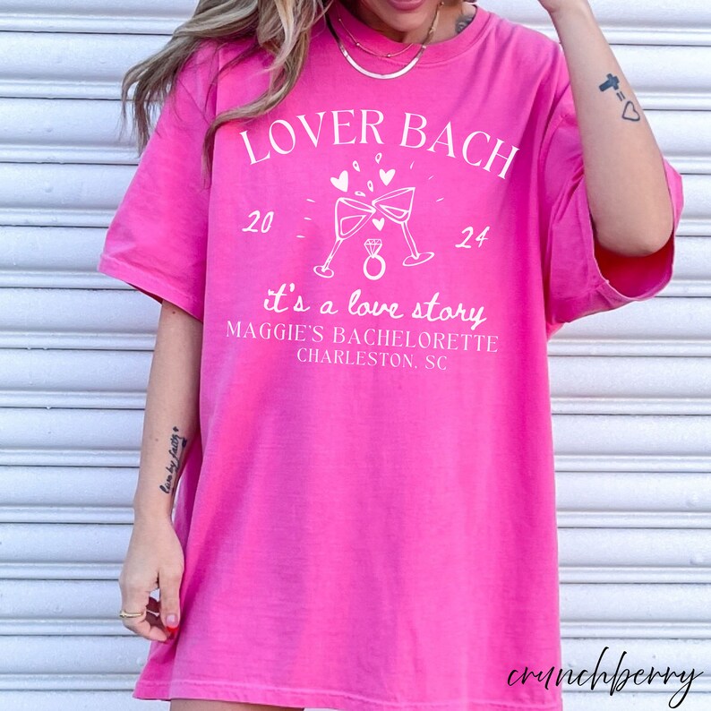 May include: Pink t-shirt with white text that reads "Lover Bach 20 - 24 it's a love story Maggie's Bachelorette Charleston, SC". The shirt features a graphic of two champagne glasses clinking with a ring in the middle.