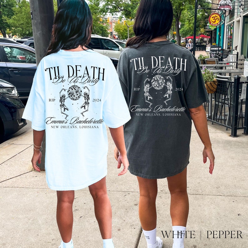 May include: Two women wearing light blue and dark gray t-shirts with a graphic print of skeletons dancing with a disco ball. The text on the shirts reads "Til Death Do Us Party" with the dates "RIP" and "2024" and the words "Emma's Bachelorette" and "New Orleans, Louisiana".