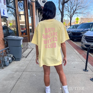 Puede incluir: Una camiseta amarilla con un gr&aacute;fico rosa que dice "Anti Social Wives Club" en la espalda.