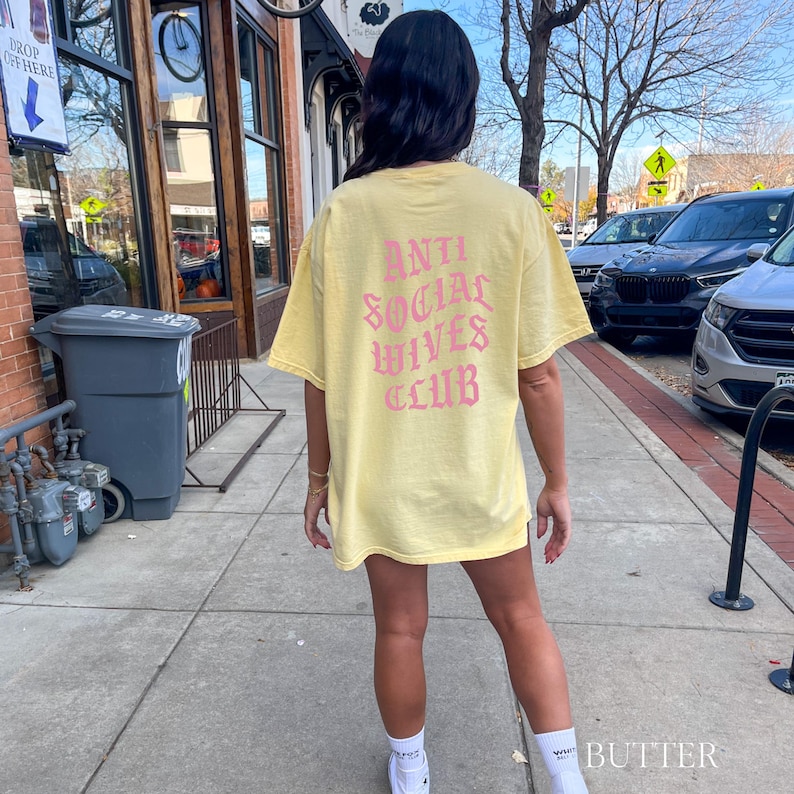 Puede incluir: Una camiseta amarilla con un gr&aacute;fico rosa que dice "Anti Social Wives Club" en la espalda.
