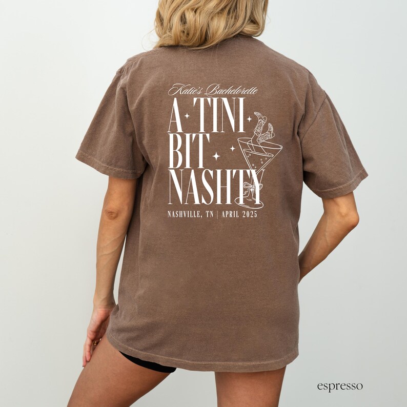 May include: A brown t-shirt with white text that reads "Katie's Bachelorette A TINI BIT NASTY Nashville, TN | April 2025". The text is surrounded by stars and a martini glass with a straw.