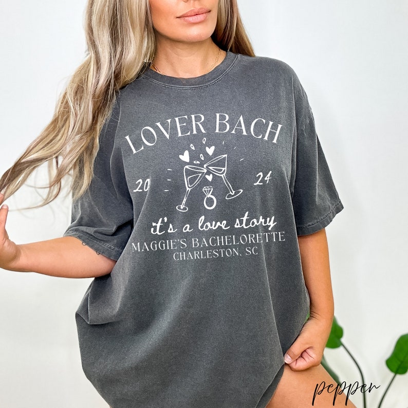 May include: A gray t-shirt with the text "Lover Bach 20 - 24 it's a love story Maggie's Bachelorette Charleston, SC" printed in white. The shirt features a graphic of two champagne glasses clinking with a ring in between.