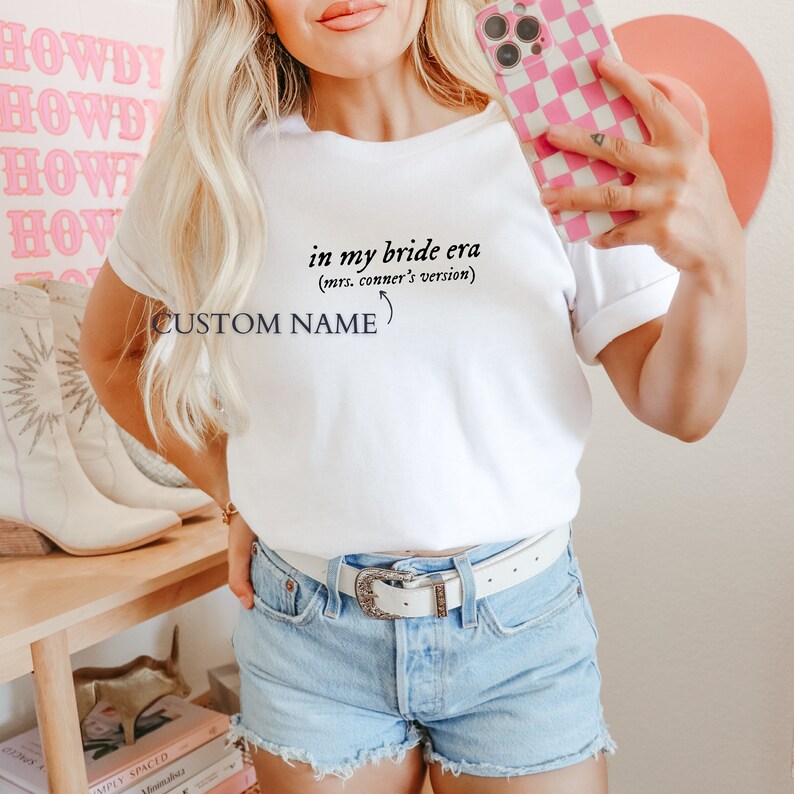 K&ouml;nnte beinhalten: Wei&szlig;es T-Shirt mit dem Text "in my bride era (mrs. conner's version)" und "CUSTOM NAME" darauf gedruckt.