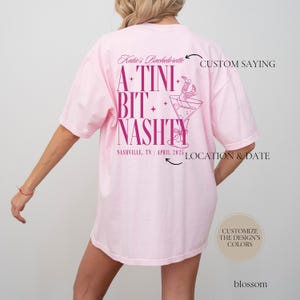 May include: Pink t-shirt with a pink graphic design that says "A-Tini Bit+ Nashty" with a martini glass and a woman's legs in cowboy boots hanging out of the glass. The text "Katie's Bachelorette" is at the top of the design. The text "Nashville, TN | April 2025" is at the bottom of the design.
