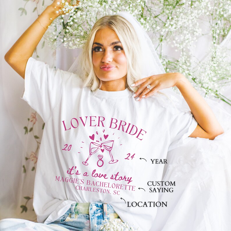May include: White t-shirt with pink text that reads "Lover Bride" with a graphic of two champagne glasses clinking and a ring. The text "It's a love story" is below the graphic. The text "Maggie's Bachelorette Charleston, SC" is below the text "It's a love story".