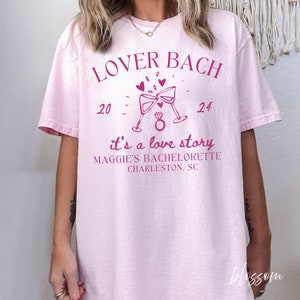May include: Pink t-shirt with the text "Lover Bach" and "it's a love story" with a graphic of two champagne glasses clinking and a ring. The text also includes the dates "20" and "24" and the words "Maggie's Bachelorette Charleston, SC".