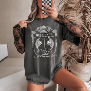 Op de afbeelding: Donkergrijs t-shirt met een wit grafisch ontwerp. Het ontwerp bevat de tekst "HIGH LADY OF THE NIGHT COURT" en "MACY'S BACHELORETTE". Het shirt heeft een ronde hals en korte mouwen. Het shirt is gemaakt van een zacht, comfortabel materiaal.