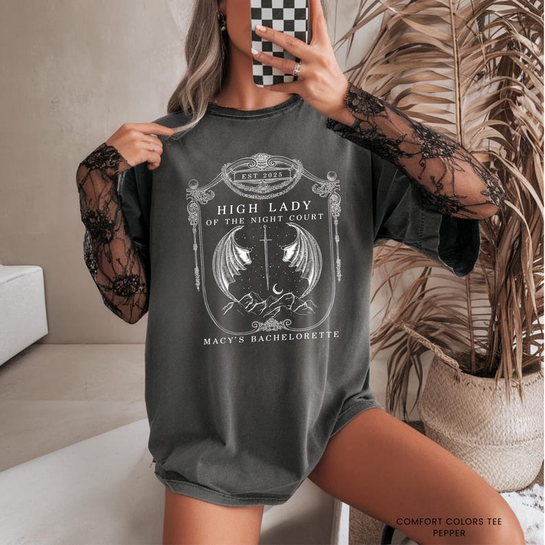 Op de afbeelding: Donkergrijs t-shirt met een wit grafisch ontwerp. Het ontwerp bevat de tekst "HIGH LADY OF THE NIGHT COURT" en "MACY'S BACHELORETTE". Het shirt heeft een ronde hals en korte mouwen. Het shirt is gemaakt van een zacht, comfortabel materiaal.