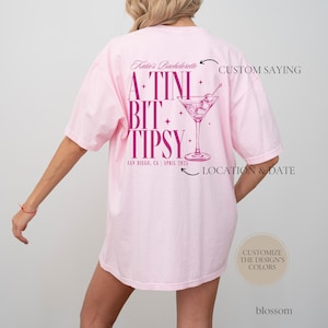 May include: Pink t-shirt with a pink martini glass graphic and the text "A-TINI BIT TIPSY" with the words "Katie's Bachelorette" and "SAN DIEGO, CA APRIL 2025" below the graphic. The text "CUSTOM SAYING" and "LOCATION & DATE" are written above and below the graphic, respectively.