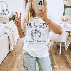 K&ouml;nnte beinhalten: Wei&szlig;es T-Shirt mit dem Text "Til Death Do Us Party" &uuml;ber einer Grafik von Skeletten, die um eine Discokugel tanzen. Der Text "Emma's Bachelorette, New Orleans, Louisiana" steht darunter. Das Jahr 2024 ist ebenfalls aufgedruckt.