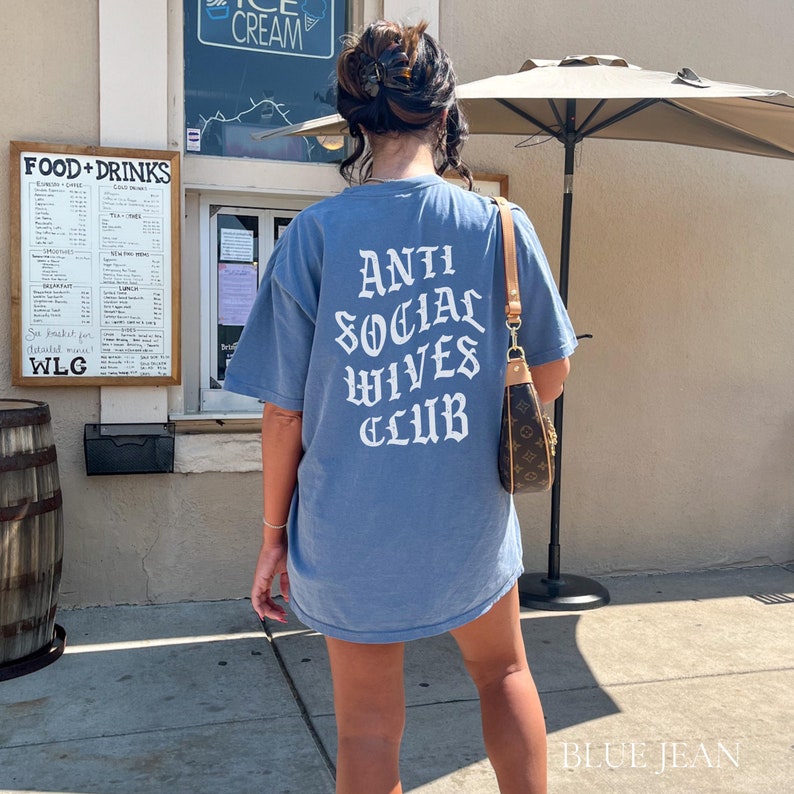 Puede incluir: Una camiseta azul con el texto "Anti Social Wives Club" impreso en blanco en la espalda. La camiseta la lleva puesta una persona que est&aacute; de pie frente a un edificio.