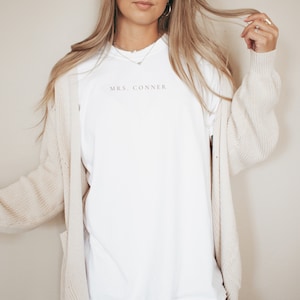 Op de afbeelding: Een wit T-shirt met de tekst "MRS. CONNER" gedrukt op de voorkant. Het shirt wordt gedragen door een persoon met lang blond haar en een beige cardigan.