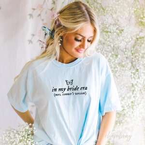 Op de afbeelding: Een lichtblauw t-shirt met een vlinder afbeelding en de tekst "in my bride era (mrs. conner's version)"