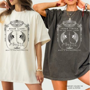 Op de afbeelding: Twee oversized T-shirts, &eacute;&eacute;n ivoor en &eacute;&eacute;n peper, met een gedetailleerde afbeelding. De afbeelding bevat de tekst "HIGH LADY OF THE NIGHT COURT" en "INNER CIRCLE OF THE NIGHT COURT", met "EST 2025" erboven en "MACY'S BACHELORETTE" eronder. De shirts zijn gelabeld "COMFORT COLORS TEE IVORY & PEPPER".