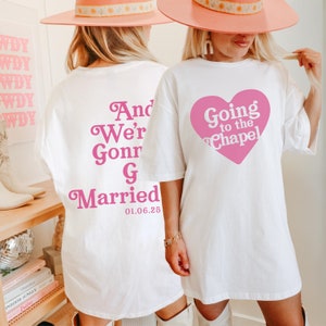 Könnte beinhalten: Zwei weiße T-Shirts mit rosa Schrift. Das vordere Shirt sagt "Going to the Chapel" mit einem rosa Herzen. Das hintere Shirt sagt "And We're Gonna Get Married 01.06.25".