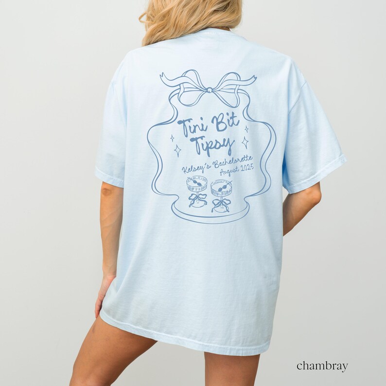 May include: Light blue t-shirt with a graphic of a blue and white cake with the text "Tini Bit Tipsy" and "Kelsey's Bachelorette August 2025".