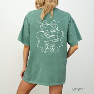 May include: A light green t-shirt with a white graphic design that says "Tini Bit Tipsy" and includes a drawing of two martini glasses. The text below the glasses says "Kelsey's Bachelorette August 2025".